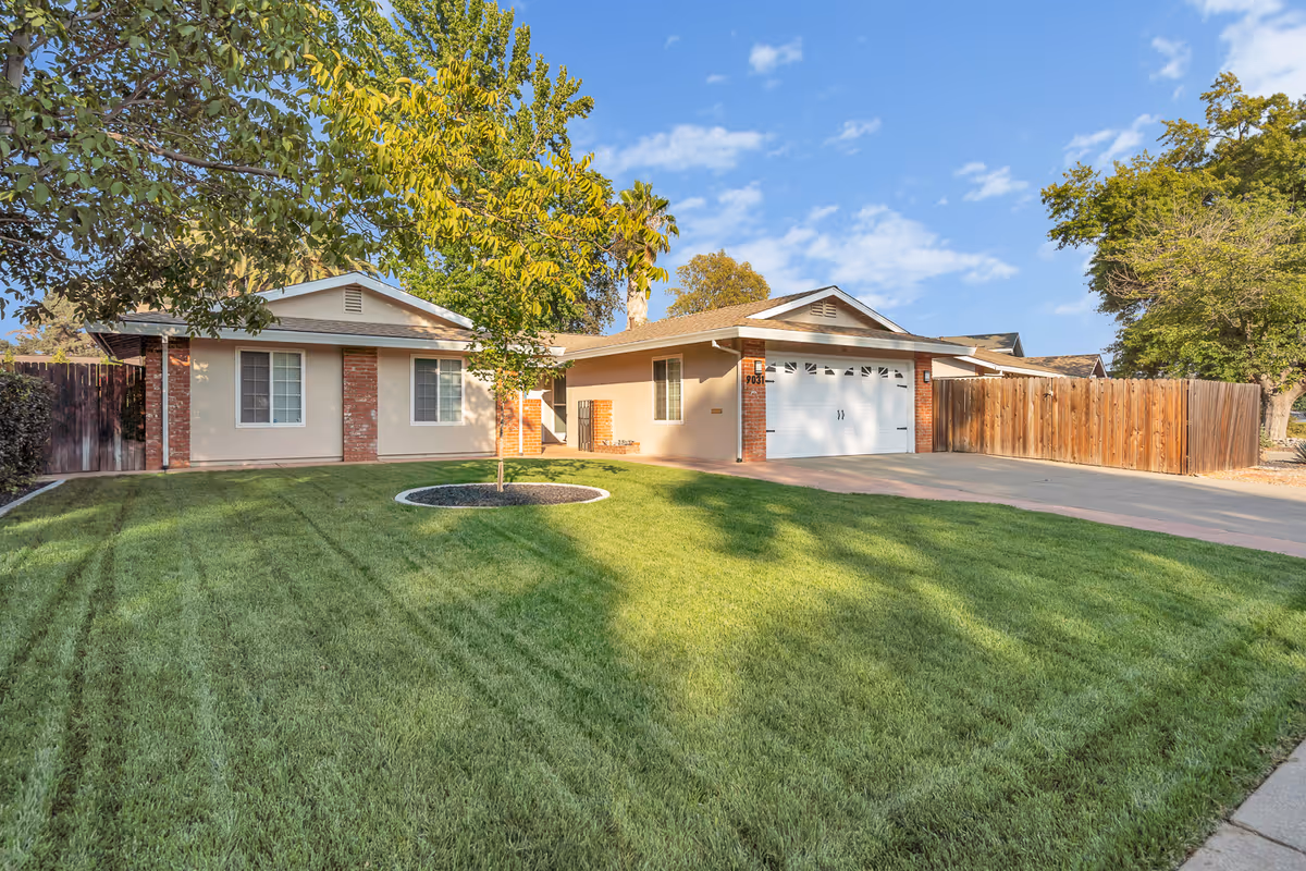 Single-story residential building with a well-maintained green lawn, a small tree in the center, a driveway leading to a white garage door, and a wooden fence on the right side under a partly cloudy blue sky.