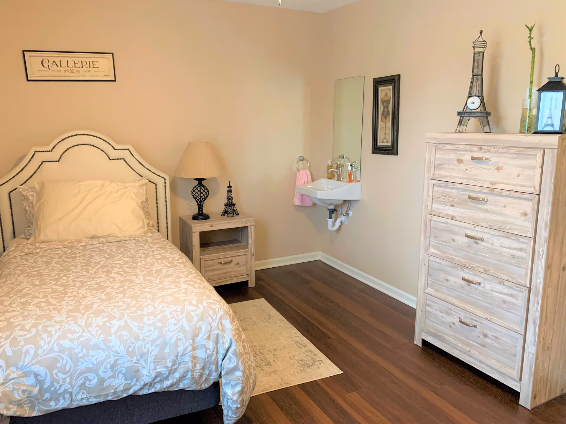 A cozy bedroom with a single bed featuring a patterned beige and white comforter and a beige pillow. Next to the bed is a wooden nightstand with a black lamp and a small Eiffel Tower decoration. On the right wall, there is a small white sink with a pink hand towel hanging on a ring, a mirror above it, and a framed picture. To the right of the sink is a wooden dresser with five drawers, decorated with a small Eiffel Tower clock, a bamboo plant, and a lantern. The room has wooden flooring and beige walls with a framed sign that reads 'GALLERIE' above the bed.