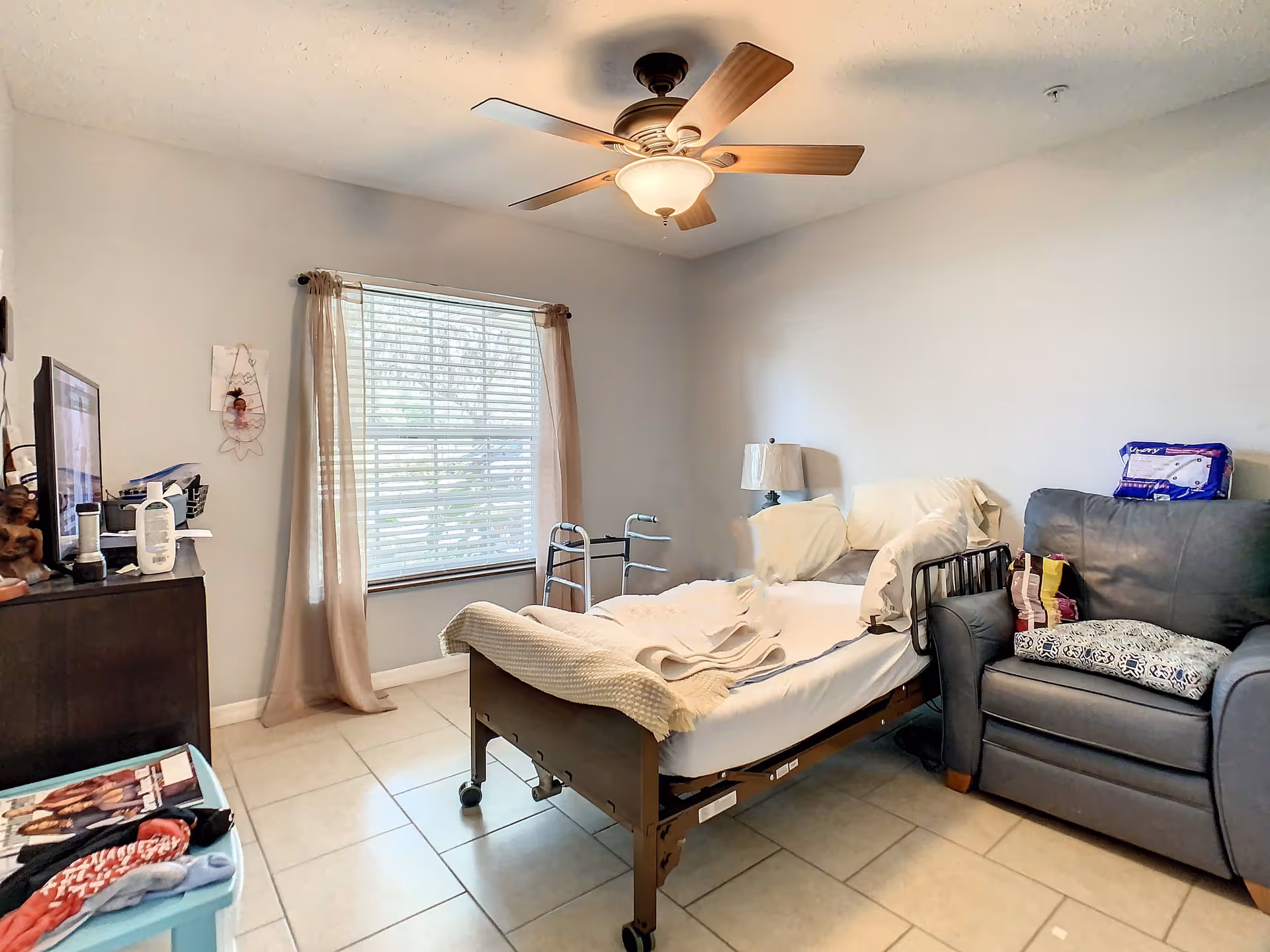 Small bedroom in a senior facility with an adjustable bed, recliner, dresser, window with curtains, ceiling fan and a walker.