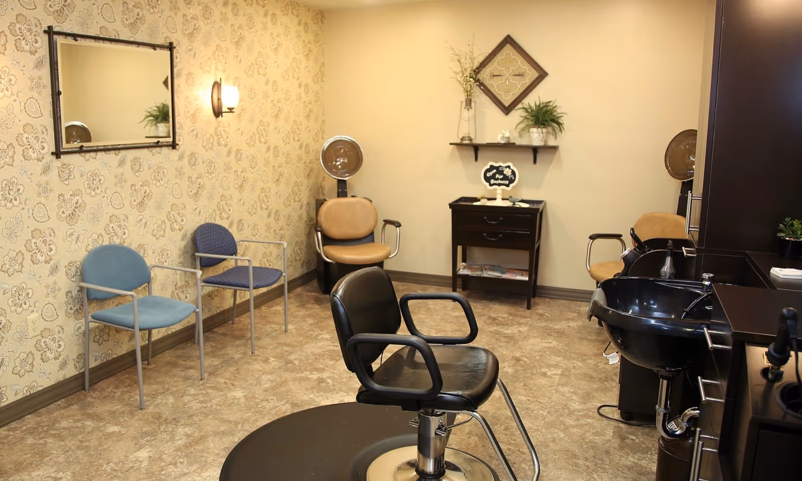 Interior of a hair salon area with a black styling chair in the center, two beige salon chairs with hair dryers in the back, two waiting chairs against a floral wallpapered wall, a black sink for washing hair, and a small cabinet with plants and decor on the wall.