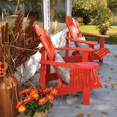 Two red Adirondack chairs with colorful patterned cushions sit on a concrete patio outside a building with large windows. There are autumn leaves scattered on the ground and a pot with orange flowers and dried plants nearby. Green trees and grass are visible in the background.