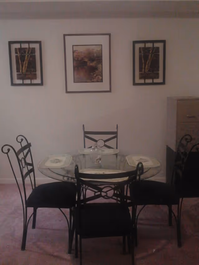 A dining area with a round glass table and four black metal chairs with cushioned seats. The table has four placemats and a small decorative item in the center. On the wall behind the table, there are three framed pictures with nature-themed artwork. To the right, there is a filing cabinet.