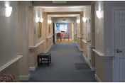 A long, well-lit hallway in a skilled nursing facility with beige walls, blue carpet, wall-mounted lights, and handrails on both sides. There is a small bench on the left side and a door on the right. At the end of the hallway, a dining area with tables and chairs is visible.