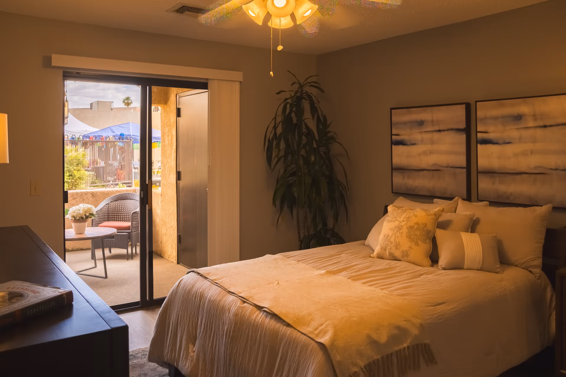 A cozy bedroom with a neatly made bed featuring multiple pillows and a beige blanket. Two abstract paintings hang on the wall above the bed. A large potted plant is in the corner near a sliding glass door that opens to a small outdoor patio with a table, chairs, and a potted plant. A ceiling fan with lights is on the ceiling, and a dresser with a book on it is visible on the left side.