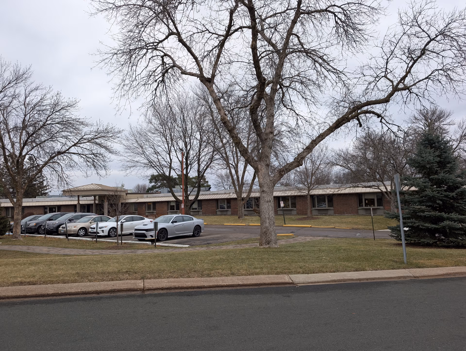 Exterior view of a single-story brick building with a metal roof, surrounded by leafless trees and a grassy area. Several cars are parked in a parking lot in front of the building under an overcast sky.