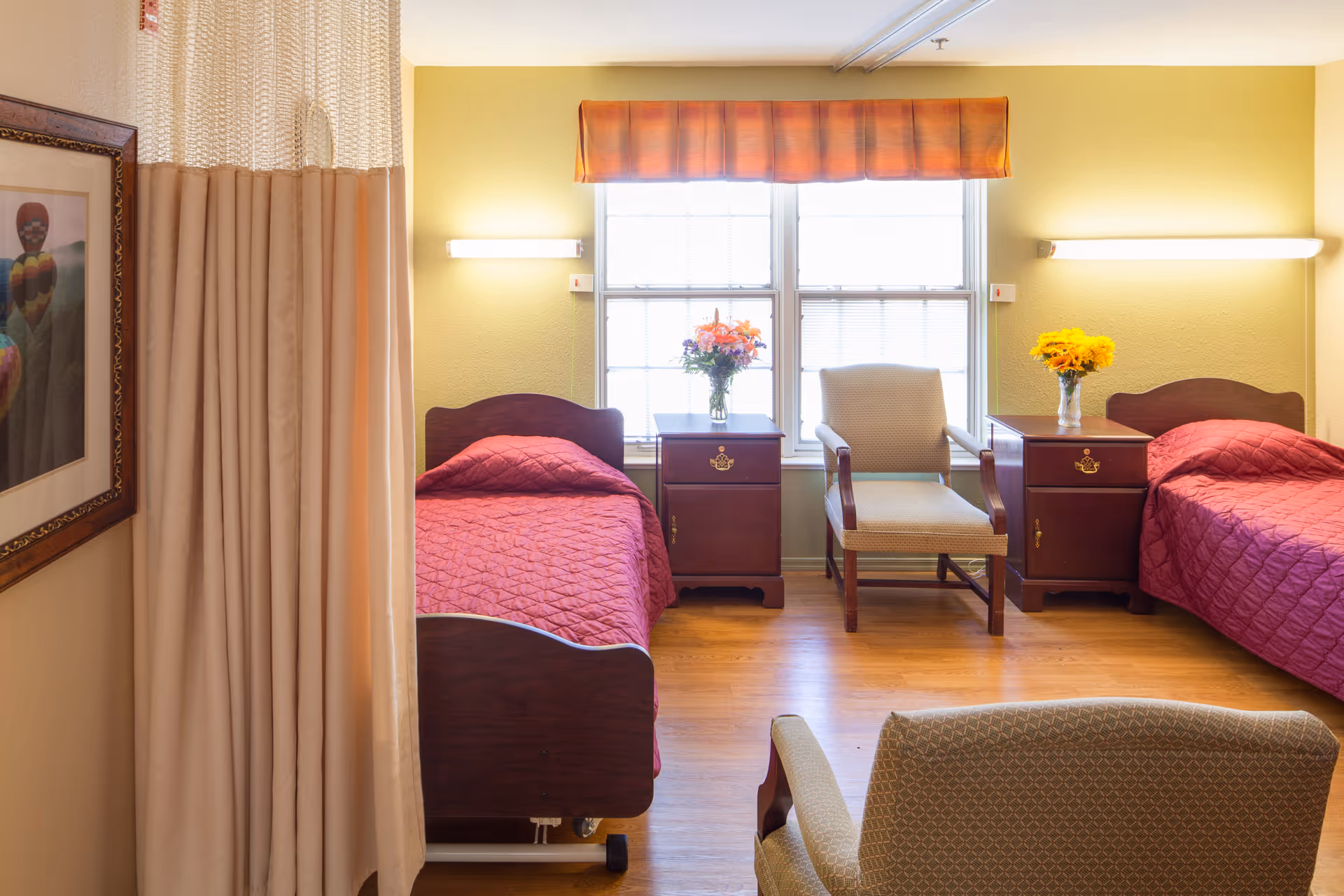 A bright room with two single beds covered in red quilts, each with a wooden headboard. Between the beds are two wooden nightstands, each holding a vase of flowers. A beige armchair is positioned between the nightstands in front of a large window with a valance. A privacy curtain is partially visible on the left side, and a framed picture hangs on the wall. The floor is wooden, and the walls are painted light yellow.