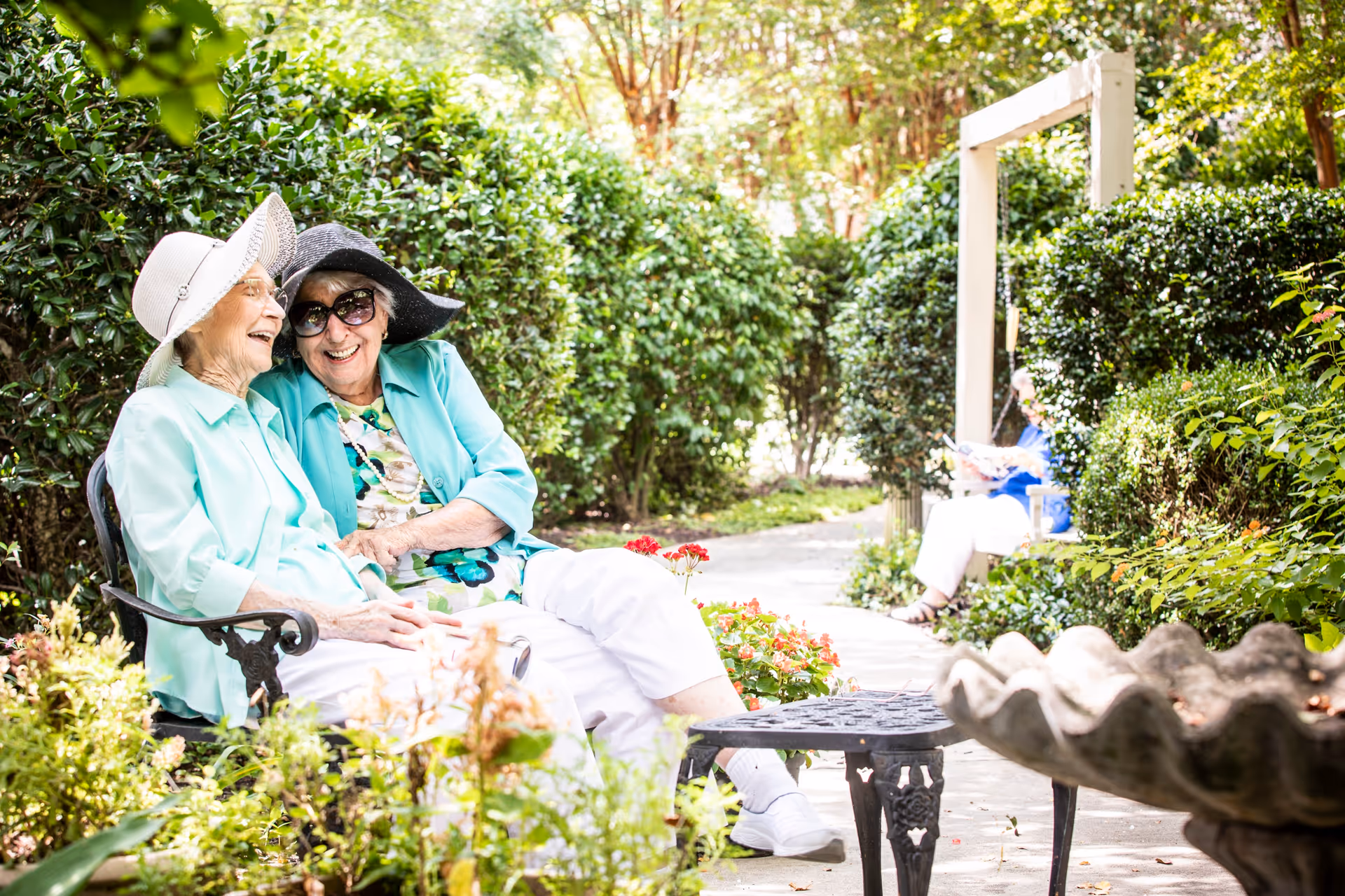 Two elderly women wearing sun hats and light-colored clothing sitting on a bench in a lush garden, smiling and enjoying each other's company. In the background, another person is sitting on a bench reading, surrounded by greenery and flowers.