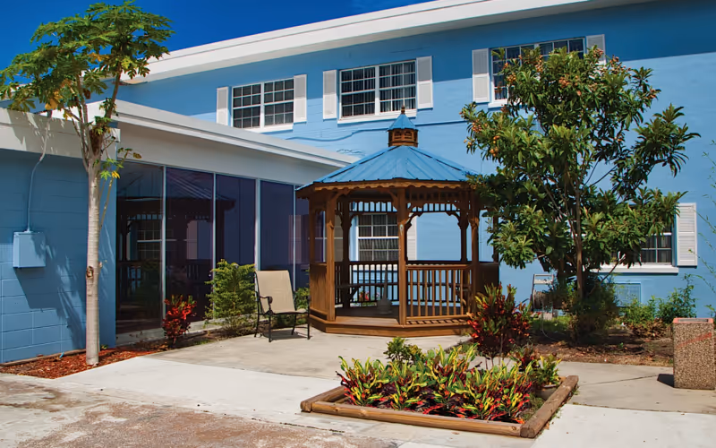 A sunny courtyard with a wooden gazebo, landscaping, a chair, and the blue two-story building exterior.