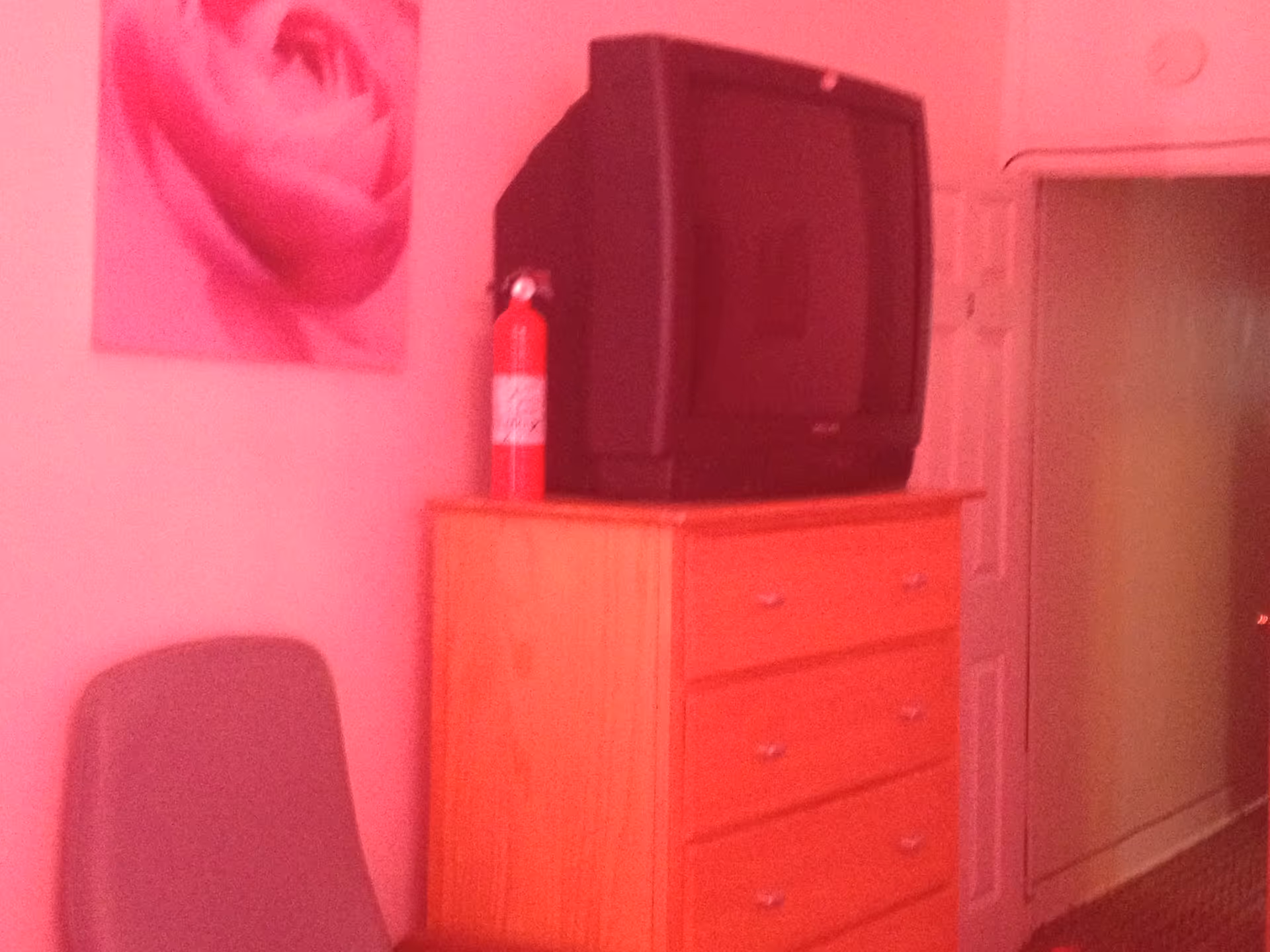 A bedroom corner with a wooden dresser holding an old-style CRT television and a red fire extinguisher. A gray chair is positioned next to the dresser, and a pink floral painting hangs on the wall above the chair. A door and a hallway are visible in the background.