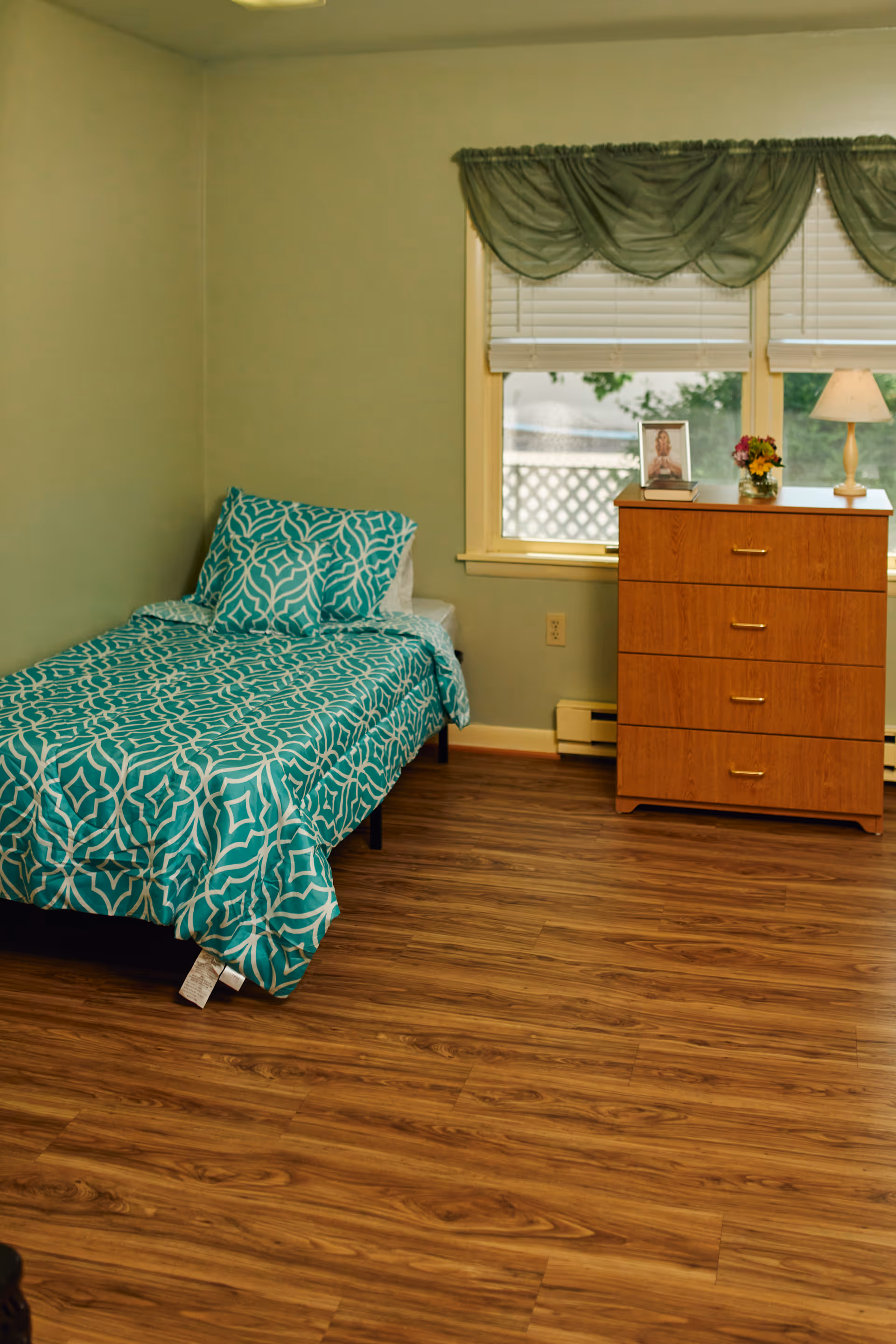 A small bedroom with a single bed covered in a teal and white patterned bedspread and matching pillow. Next to the bed is a wooden dresser with four drawers, topped with a small lamp, a framed photo, and a small flower arrangement. The room has light green walls, wood flooring, and a window with white blinds and green valance curtains.