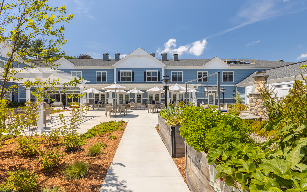 Outdoor patio area of The Branches of Framingham facility with tables, chairs, and umbrellas set up on a concrete surface. There are garden beds with green plants and shrubs on either side of the walkway leading to the building. The building is two stories with blue siding and white trim under a clear blue sky.