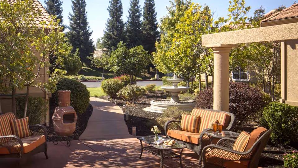 Outdoor patio area with cushioned wicker chairs and a loveseat arranged around a glass coffee table. The patio is surrounded by green shrubs, trees, and a stone fountain in the background under a clear blue sky.