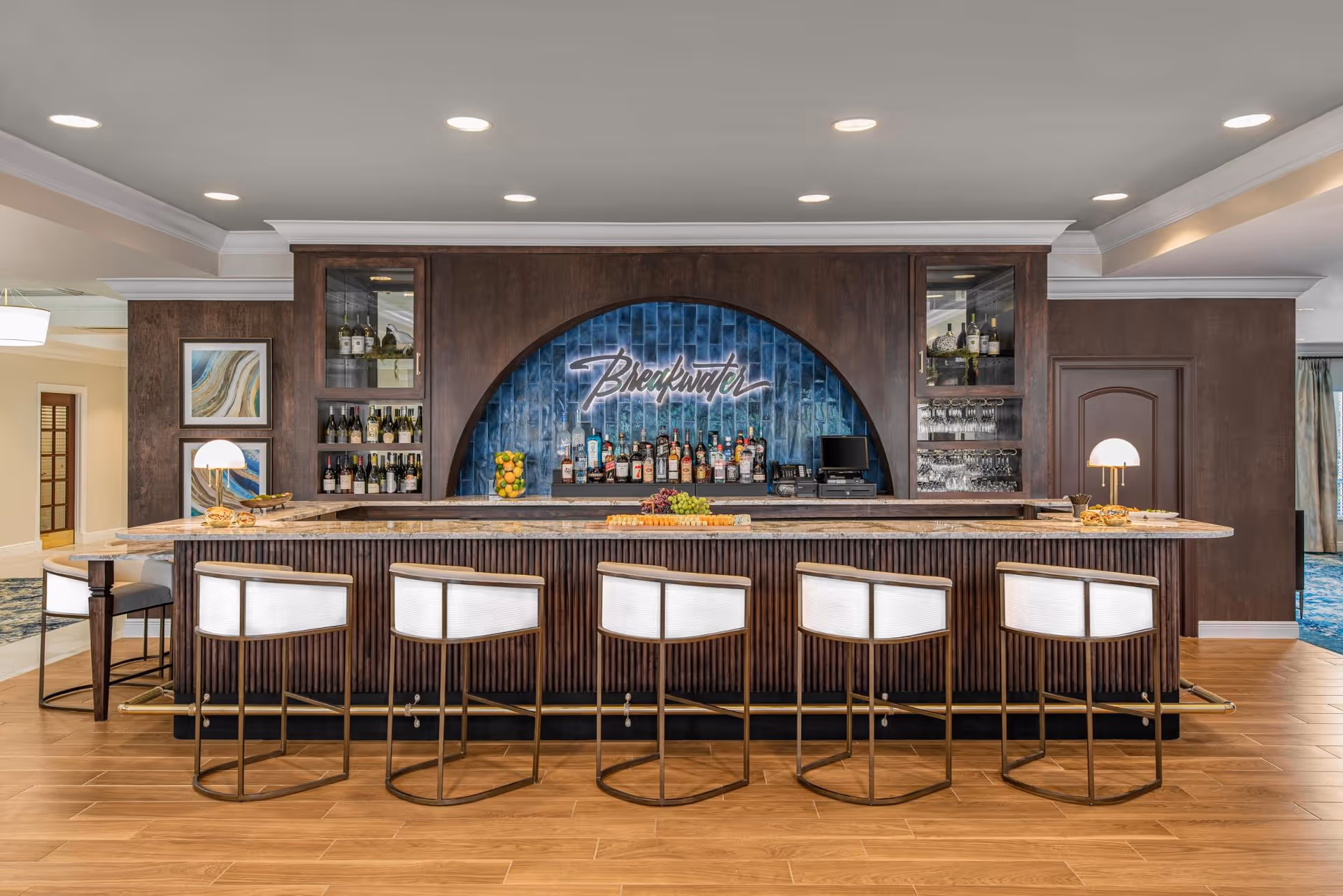 A modern bar area with six white cushioned bar stools lined up in front of a marble countertop. Behind the bar is a dark wood panel with a blue tiled arch displaying the word 'Breakwater' in neon-style lettering. Shelves on either side hold bottles of wine and glasses. The floor is wood, and the ceiling has recessed lighting.