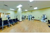 A spacious fitness room with light yellow walls and wood-patterned flooring. The room contains several exercise machines arranged around the perimeter, and a row of black chairs is lined up against one wall. The ceiling has recessed lighting and a clock is mounted on the far wall.