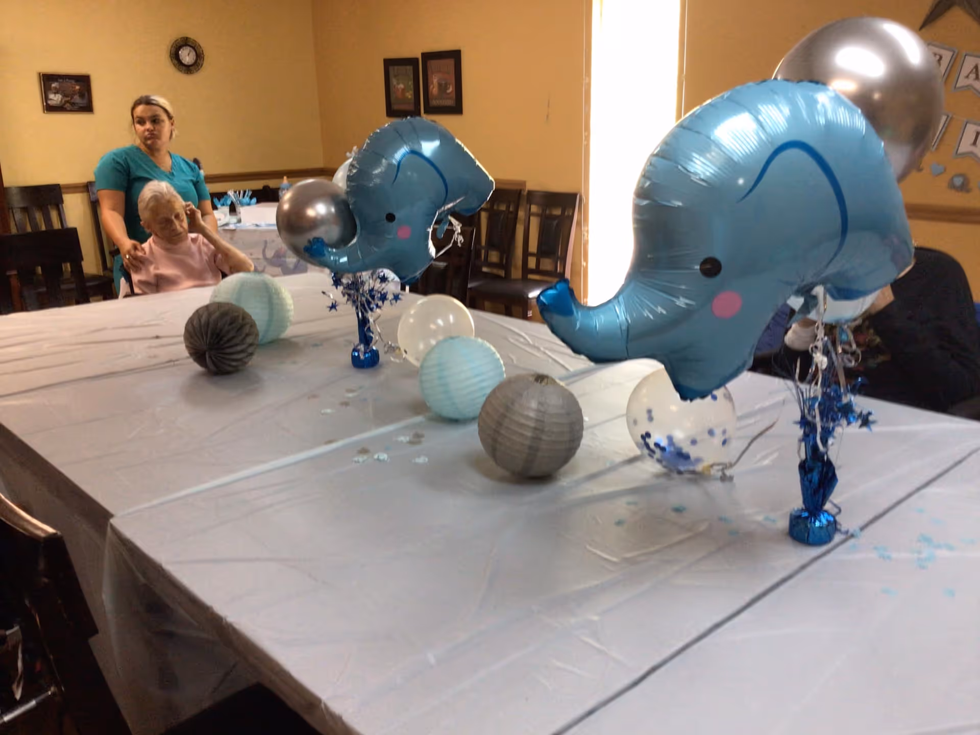 A decorated table with blue and silver balloons shaped like elephants and round paper lanterns in blue, white, and gray. In the background, a woman in teal scrubs is standing behind an elderly woman seated at the table in a room with beige walls and wooden chairs.