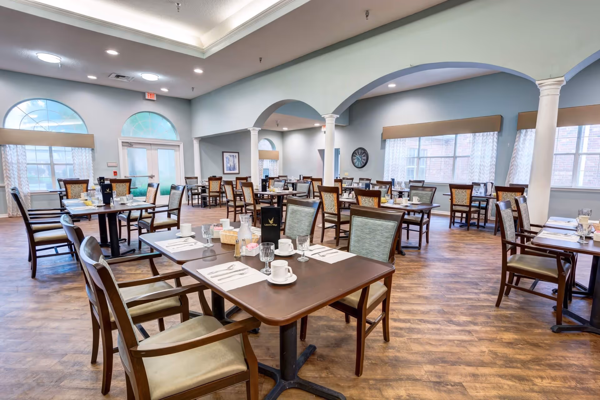 A spacious dining room with multiple wooden tables and chairs arranged neatly. Each table is set with white placemats, cups, glasses, and silverware. The room has large windows with light curtains allowing natural light to fill the space. The ceiling features recessed lighting and decorative molding, and there are white columns supporting archways within the room.