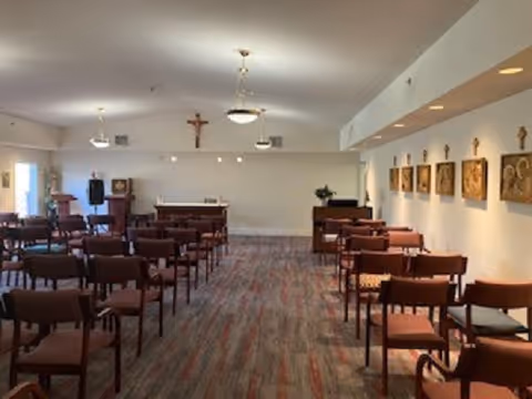 Interior view of a chapel or prayer room with rows of wooden chairs facing an altar at the front. The walls are decorated with religious icons and a crucifix is mounted above the altar. The room is well-lit with ceiling lights and has a carpeted floor.