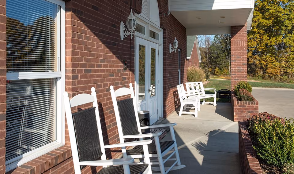 Outdoor patio area of a senior living facility with white rocking chairs and benches along a brick wall. There are two white wall-mounted lanterns beside a glass door, and some green shrubs in brick planters. Trees with autumn foliage are visible in the background under a clear sky.