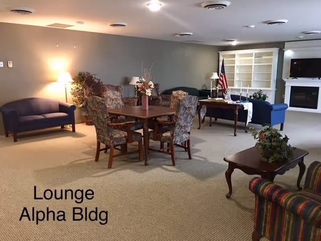A spacious lounge area with multiple seating options including floral upholstered chairs around a wooden table, a blue loveseat, and a striped armchair. The room has beige carpet, neutral-colored walls, several lamps, a fireplace with a TV above it, built-in white shelves, and an American flag in the corner.