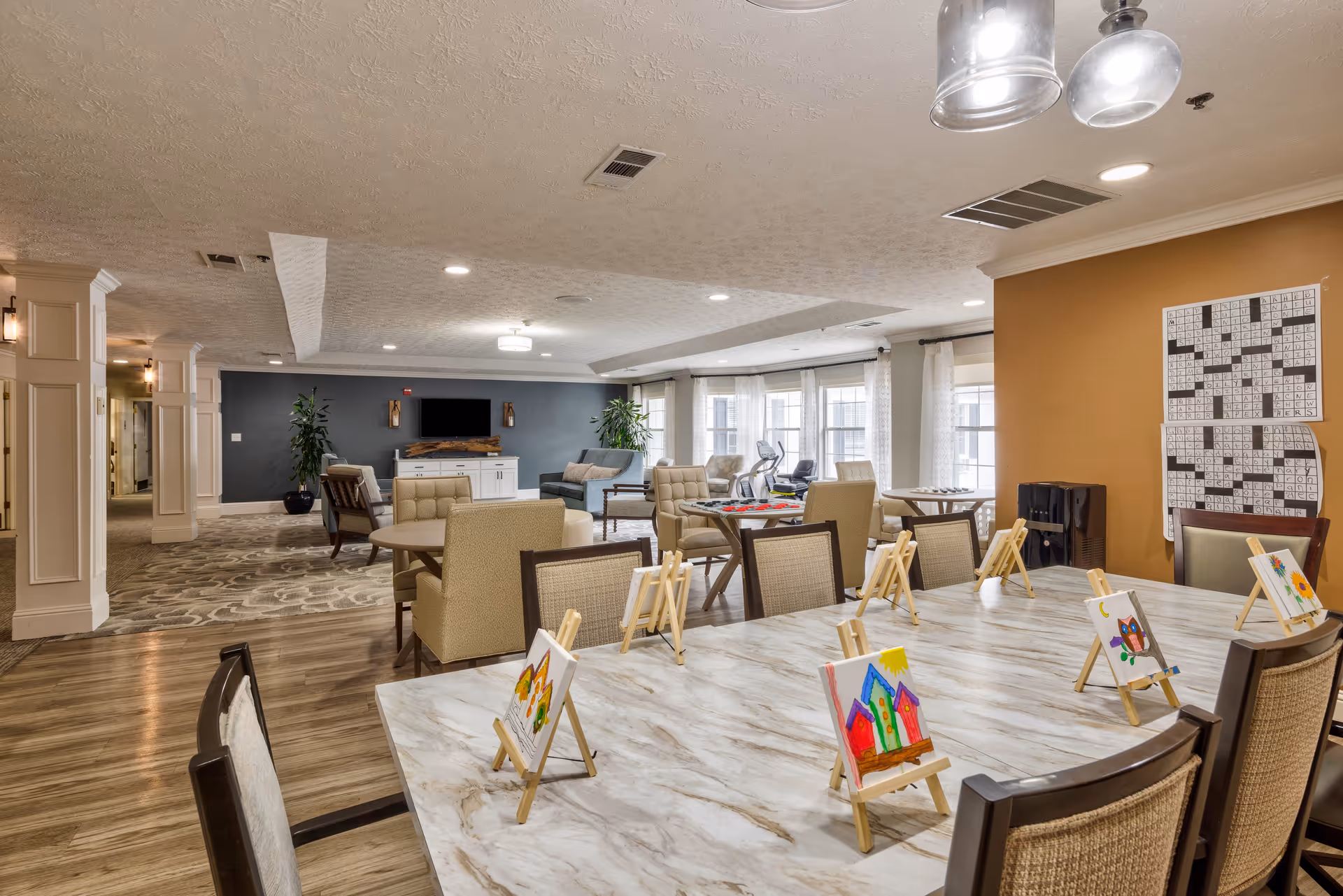 A spacious, well-lit common area in a senior living facility featuring a large table with small easels holding colorful paintings, several comfortable chairs and sofas, a television mounted on a dark accent wall, large windows with sheer curtains, and two large crossword puzzles displayed on an orange wall.