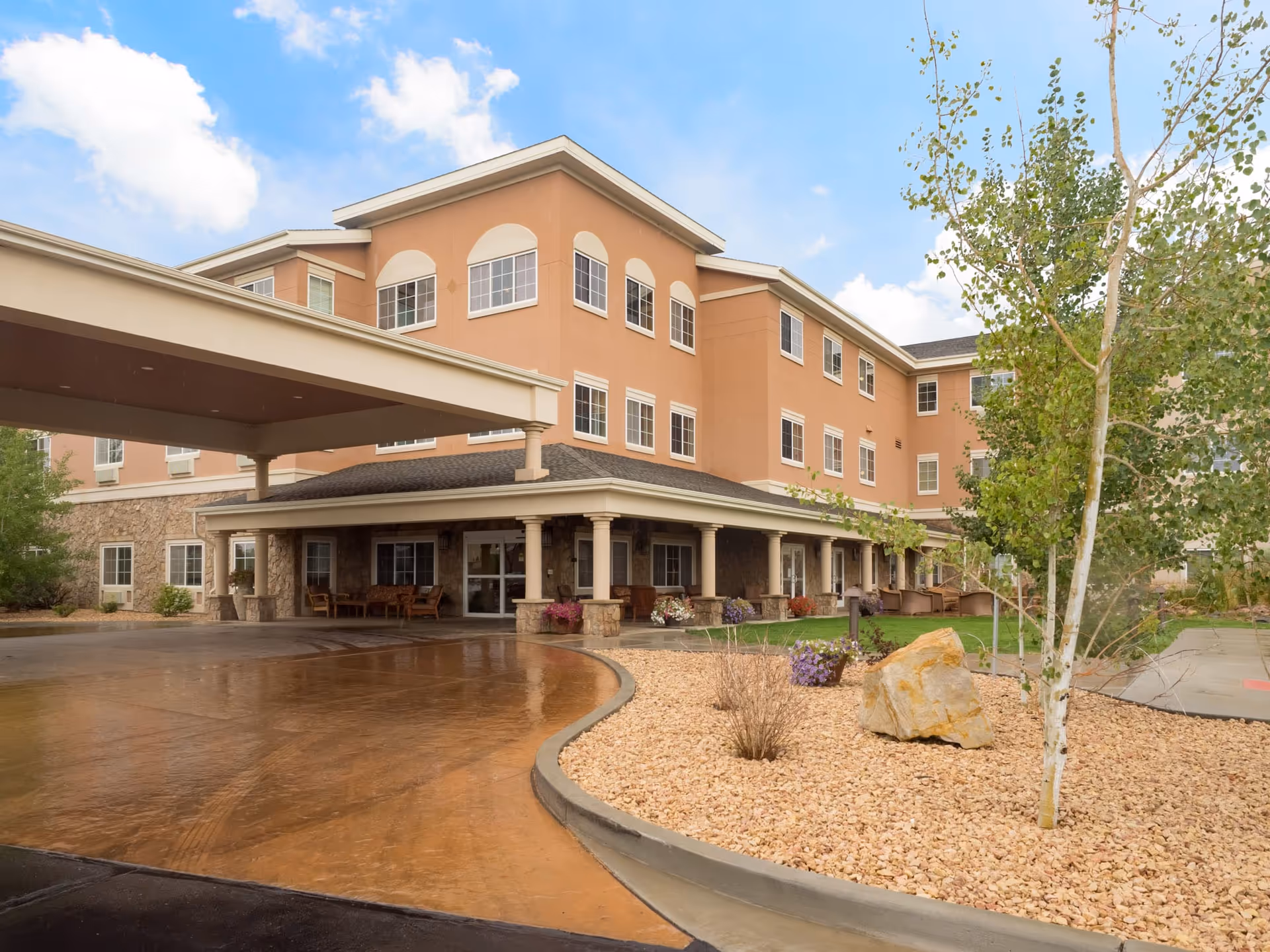 Exterior view of a three-story senior living facility building with a covered entrance driveway, stone and stucco facade, multiple windows, and landscaped area with rocks, plants, and a tree under a partly cloudy sky.