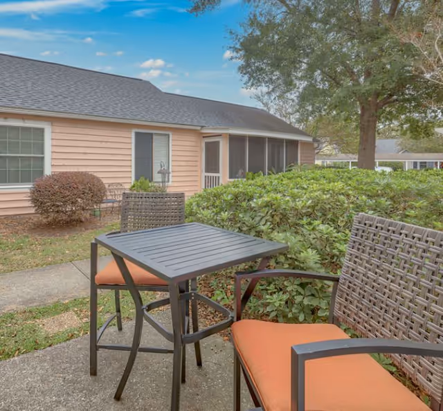 Outdoor patio area with a black metal table and two chairs with orange cushions. The patio is surrounded by green bushes and trees, with a peach-colored building and a screened porch in the background under a partly cloudy sky.