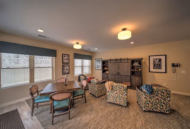 A cozy senior living common area with a wooden table and chairs on the left, and three colorful patterned armchairs arranged around a wooden cabinet with sliding barn doors on the right. The room has beige walls, carpeted flooring, two large windows with blinds and valances, framed black and white photos on the walls, and ceiling lights providing warm illumination.