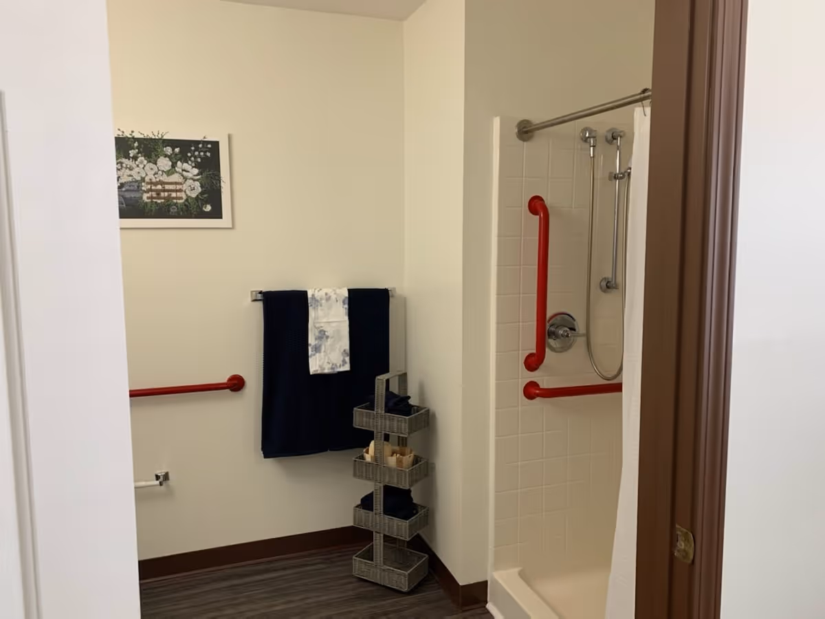 A bathroom with white walls and dark wood flooring. There is a shower with white tiles and red grab bars, a shower curtain, and a handheld showerhead. On the wall outside the shower, there is a towel rack holding two dark blue towels and one white towel with a blue pattern. A small multi-tiered wicker storage unit holds additional towels and toiletries. A framed floral picture hangs on the wall above the towel rack.