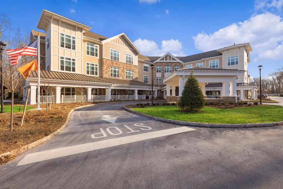 Front entrance of a three-story senior living facility with a covered porte-cochere, circular driveway, flags, and landscaped lawn under a blue sky.