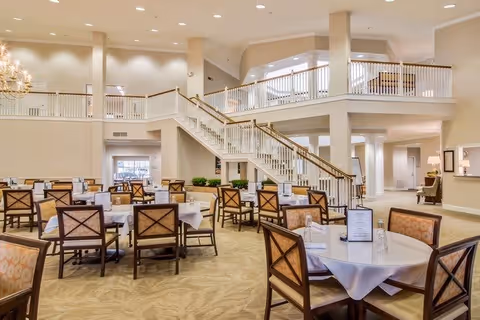 Spacious dining area with multiple round and square tables covered with white tablecloths, surrounded by wooden chairs with cushioned seats. The room features beige carpeting, cream-colored walls, and a large staircase with white railings leading to an upper level. The area is well-lit with recessed ceiling lights and has a welcoming, open atmosphere.