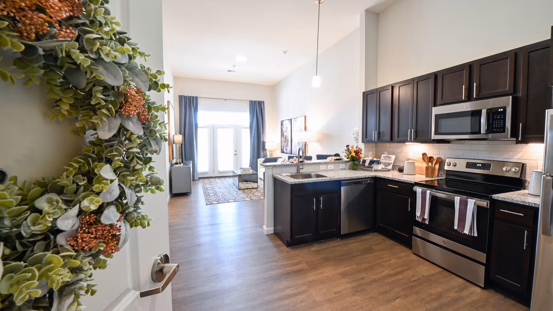 View of a modern kitchen with dark wood cabinets, stainless steel appliances including a microwave, stove, dishwasher, and refrigerator. The kitchen has granite countertops and a sink with a faucet. Beyond the kitchen is a living area with a patterned rug, a coffee table, a sofa, two lamps, and large windows with blue curtains letting in natural light. A decorative green wreath with orange berries is hanging on the door in the foreground.