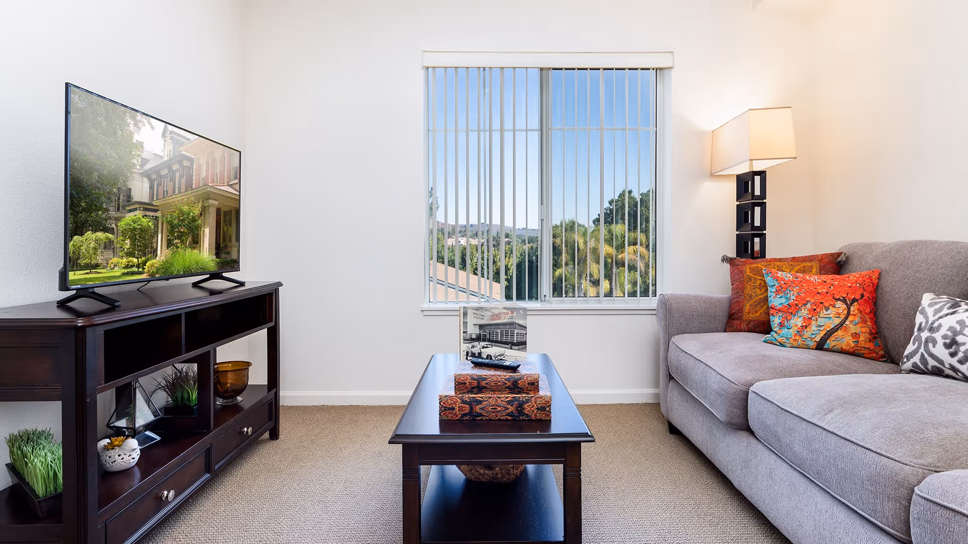 A cozy living room with a gray sofa adorned with colorful pillows, a dark wooden coffee table with decorative items, a matching TV stand with a flat-screen TV displaying an outdoor scene, and a window with vertical blinds letting in natural light.