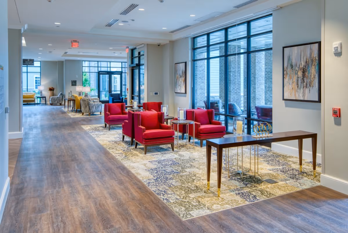 A bright and spacious interior common area with wooden flooring and large windows. The seating area features four red armchairs arranged around small tables on a patterned rug. There is a long wooden console table with decorative items, and abstract artwork hangs on the walls. The space is well-lit with natural light coming through the windows and ceiling lights.