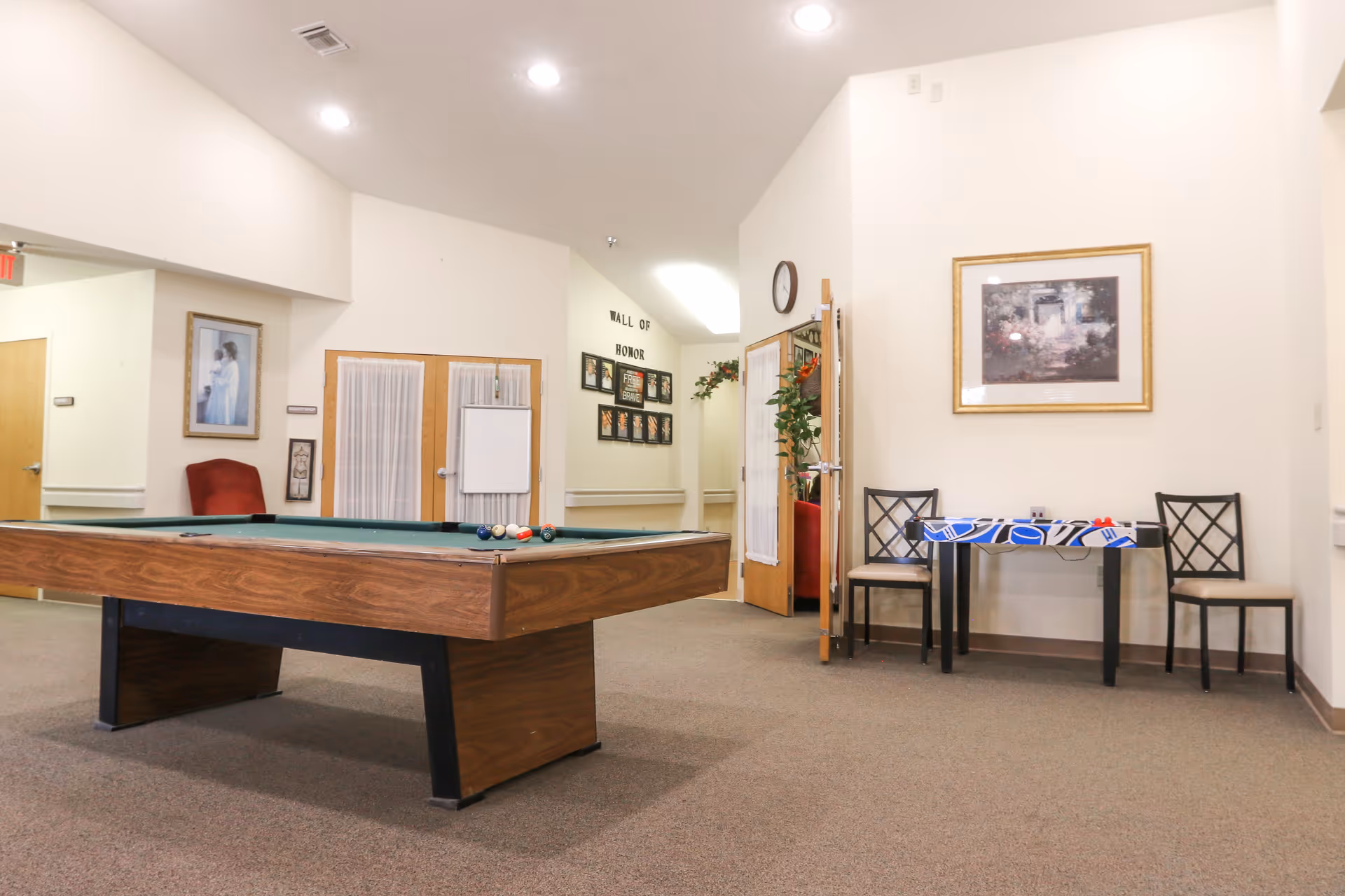 Bright common room with a pool table in the foreground, a small table and chairs against the wall, and framed pictures on the walls.