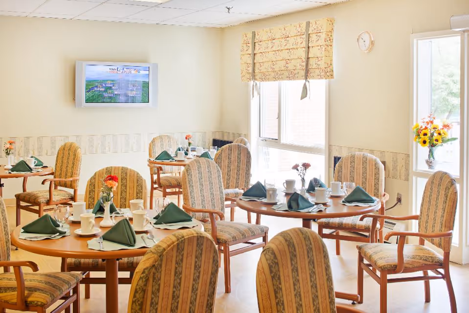 A bright dining room with several round wooden tables set with white cups, saucers, glasses, silverware, and green folded napkins. The chairs have wooden frames and striped upholstery. There are flowers in small vases on each table. A wall-mounted TV is visible, and large windows with floral-patterned blinds allow natural light to fill the room.