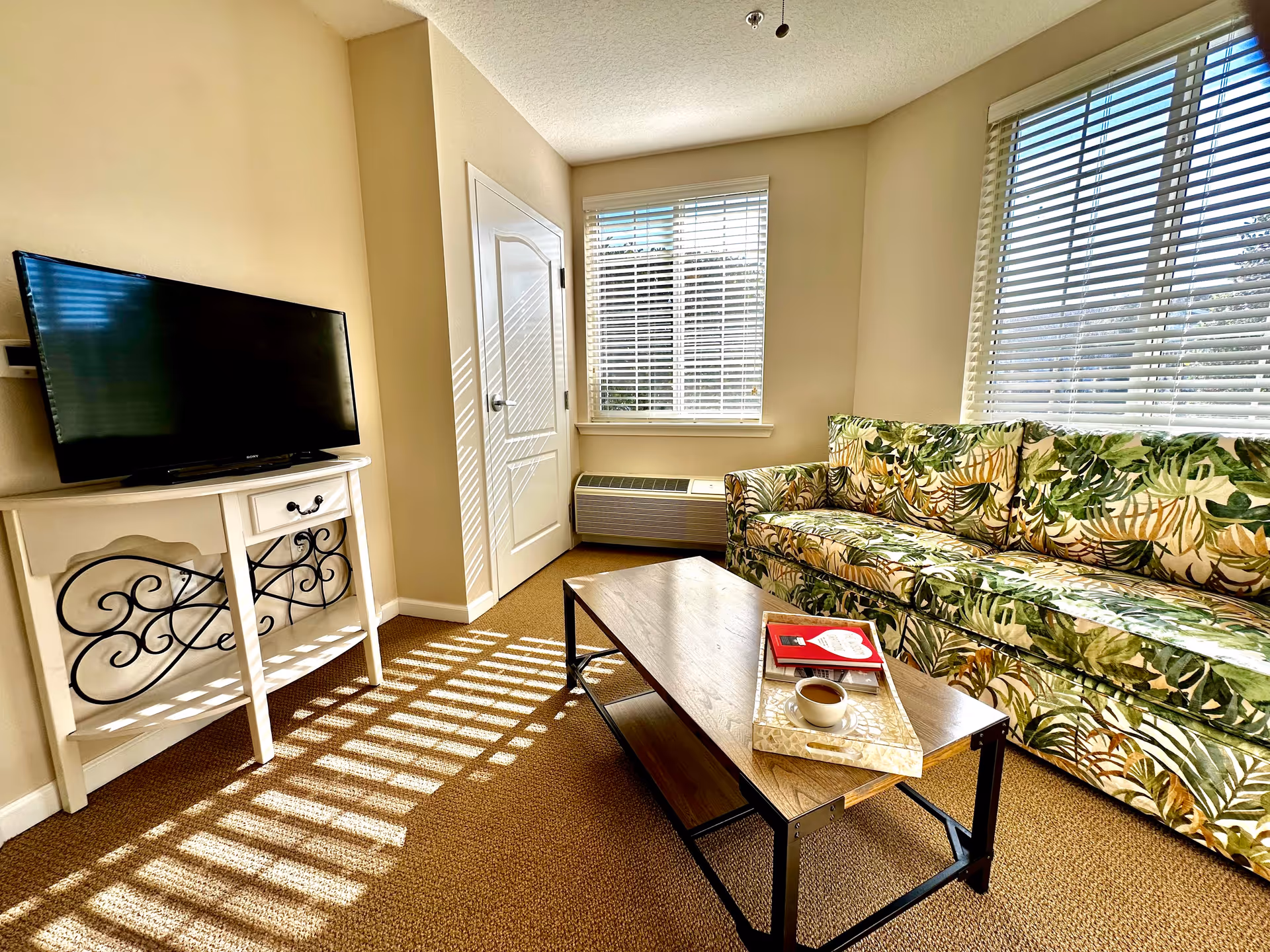A bright living room with beige walls and carpet, featuring a floral patterned sofa with green and brown leaves, a wooden coffee table with a tray holding a cup of coffee and a book, a white TV stand with decorative black metal scrollwork, and a flat-screen TV. Two windows with white blinds allow sunlight to cast striped shadows on the floor and furniture.