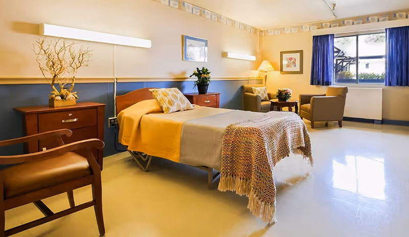 A bright, neatly arranged senior bedroom with a single bed, bedside tables, chairs and a window with blue curtains.
