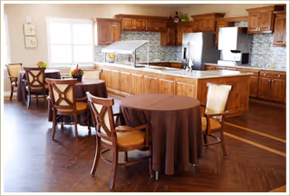 Open dining area with round tables and chairs in front of a large kitchen with wooden cabinets and an island.