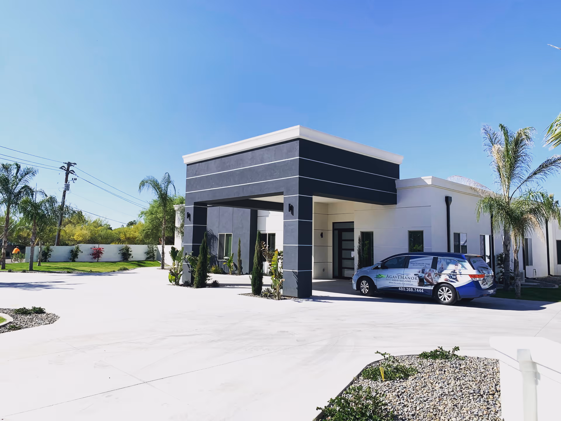 Exterior view of Agave Manor facility showing a modern building with a covered entrance, palm trees, and a branded vehicle parked in front under a clear blue sky.