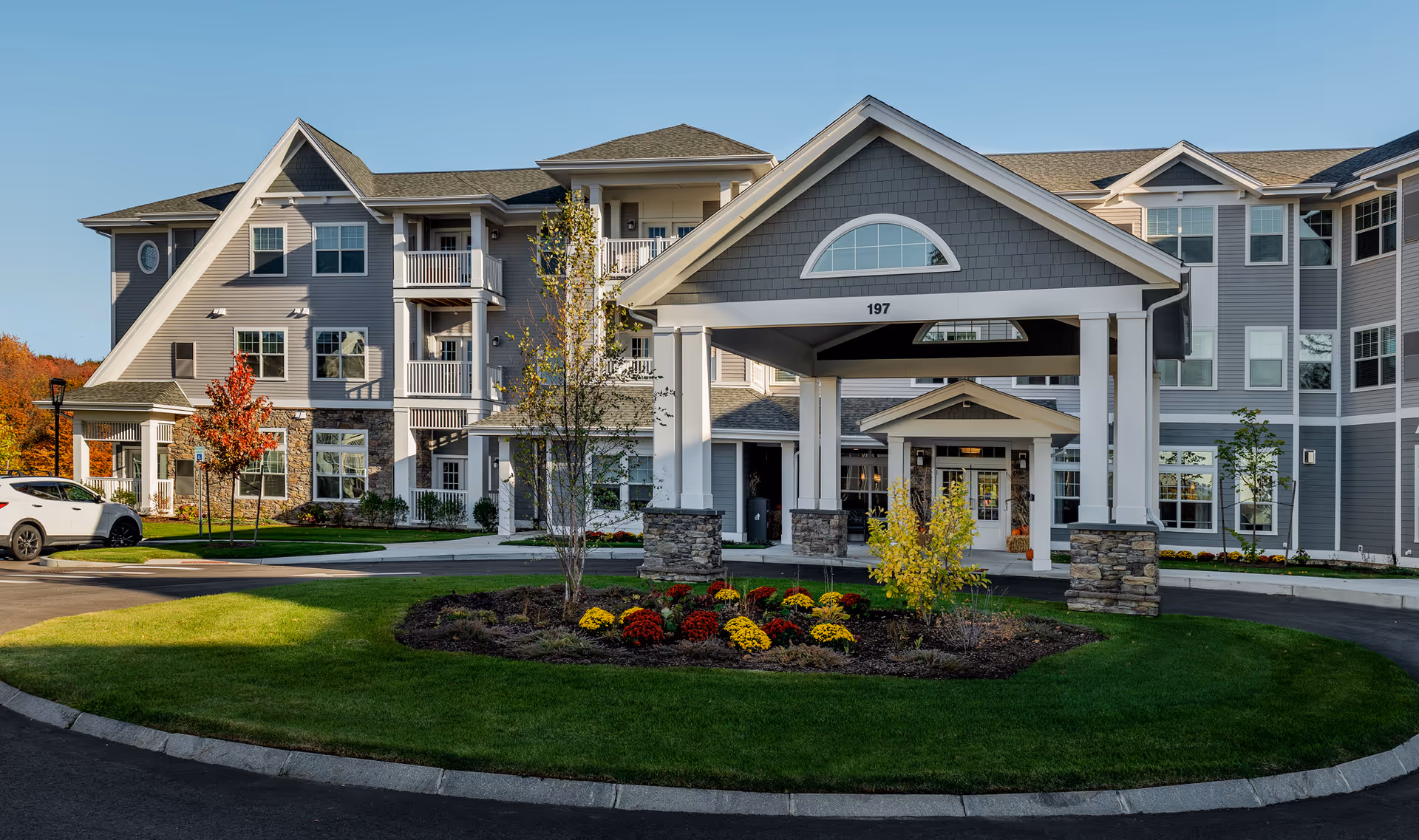 Front exterior view of The Woods - Retirement Community building with a covered entrance, well-maintained lawn, flower bed with red and yellow flowers, and a clear blue sky.