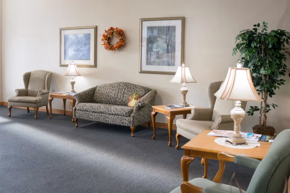 A cozy sitting area with armchairs, a patterned loveseat, side tables holding lamps and magazines, framed pictures on the wall, and a potted plant.