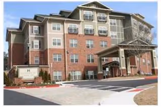 Exterior view of a multi-story senior living facility building with brick and beige siding, large windows, and a covered entrance with a driveway and parking area in front.