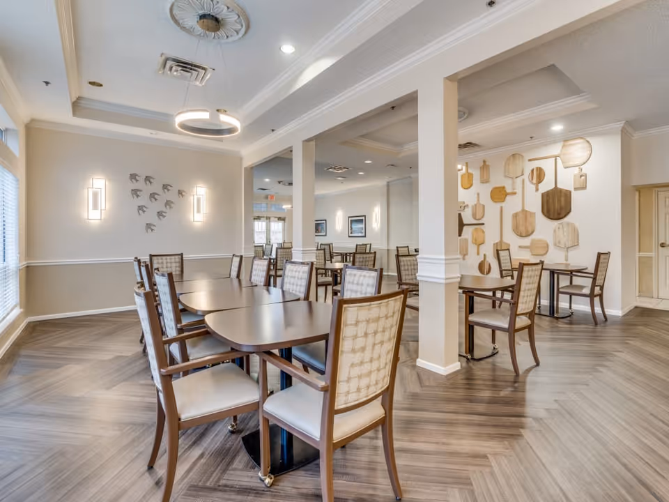 A bright and spacious dining room with multiple wooden tables and cushioned chairs arranged neatly. The room features light-colored walls with decorative wall art including a cluster of bird sculptures and various wooden paddle-shaped pieces. The floor has a herringbone pattern, and the ceiling has recessed lighting and a modern circular light fixture.