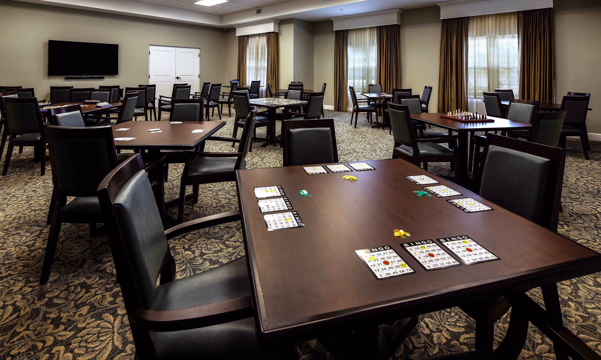 Spacious interior activity room with multiple tables and chairs set up for bingo and other games, a wall-mounted TV, and curtained windows.