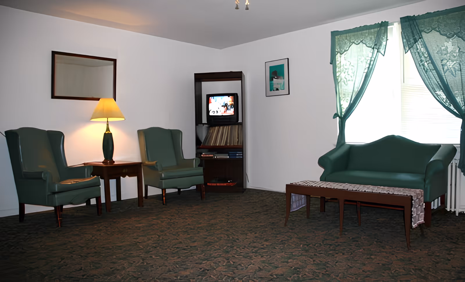 A living room with two green armchairs and a small wooden table with a lamp between them. A small green loveseat is positioned near a window with green lace curtains. A wooden coffee table with a lace runner is in front of the loveseat. A wooden shelving unit holds a small television and some books or records. The walls are white and there is a framed picture hanging on the wall.