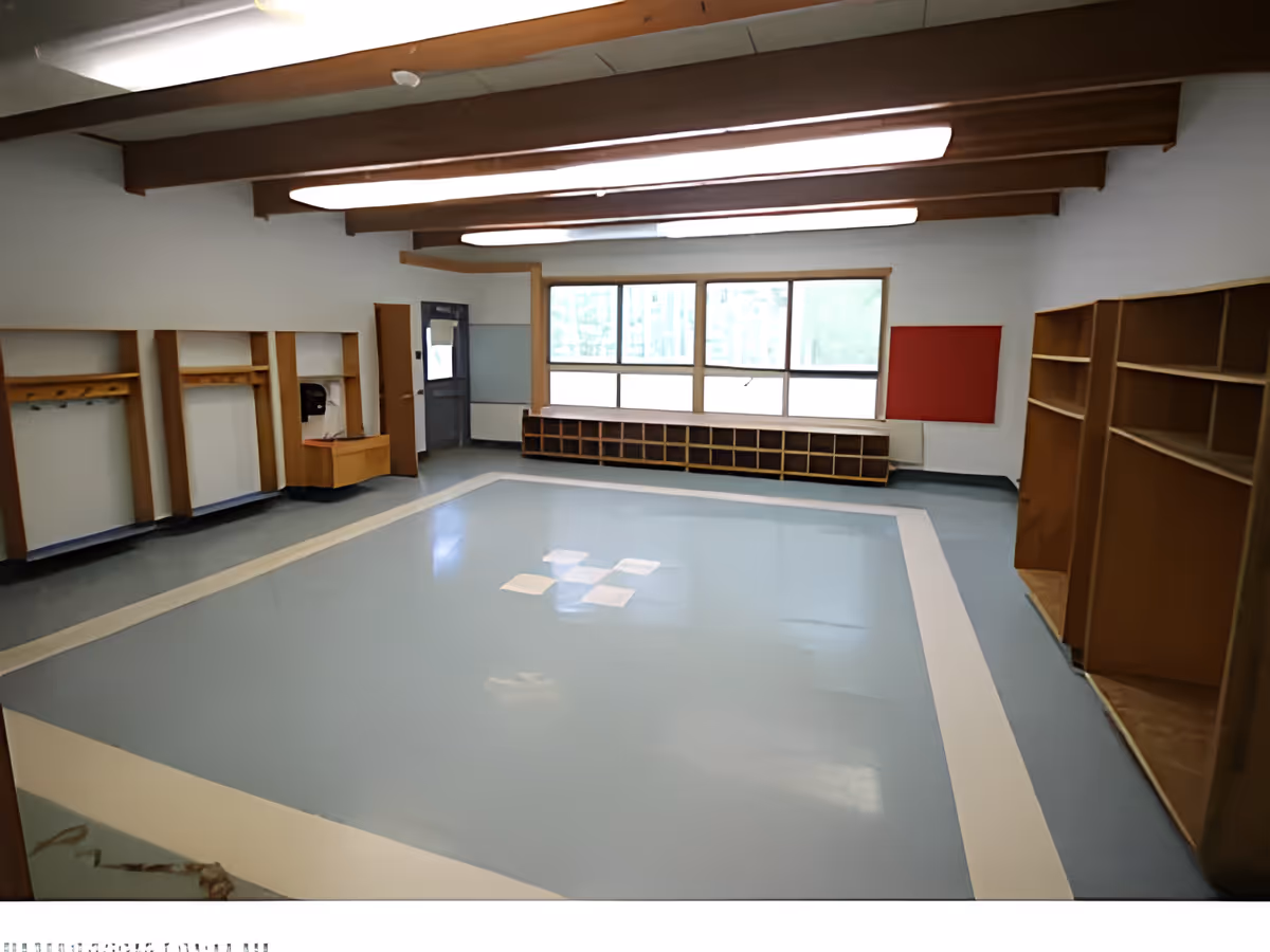Empty room with light blue and white flooring, wooden shelves and cubbies along the walls, large windows letting in natural light, and exposed wooden beams on the ceiling.