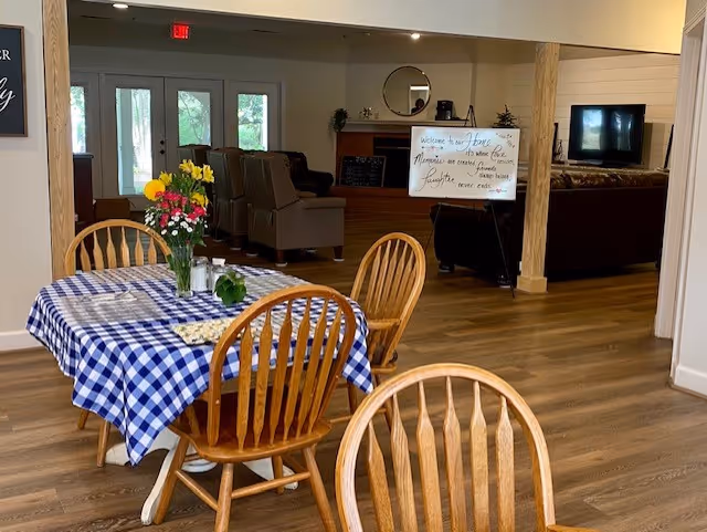 Communal dining area with a blue-and-white checkered tablecloth on a table and wooden chairs opening into a lounge with armchairs and a TV.