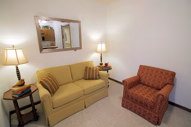 A cozy living room area with a beige sofa adorned with two striped pillows, a red patterned armchair, two wooden side tables each holding a lamp, and a large rectangular mirror on the wall reflecting part of a kitchen area.