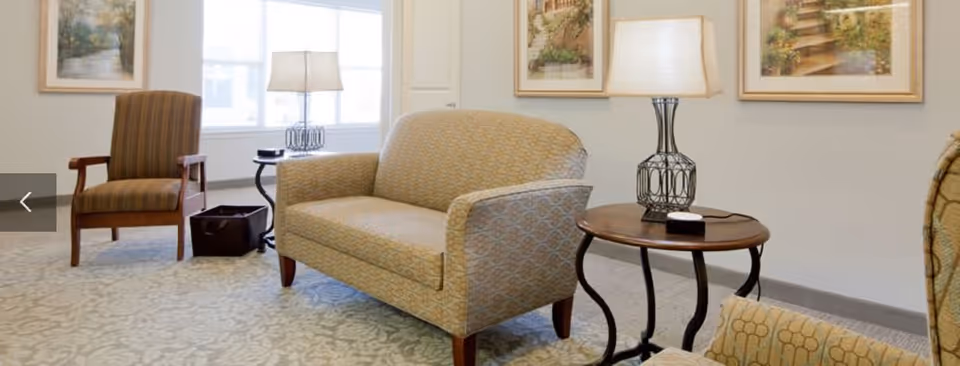A cozy seating area in a senior living facility with a patterned loveseat, a striped armchair, a round wooden side table with a decorative lamp, and framed artwork on the walls. The room is well-lit by natural light coming through a large window.