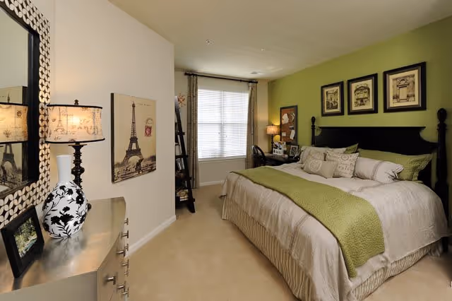 A cozy bedroom with a large bed featuring white and green bedding. The room has a green accent wall behind the bed with three framed pictures. There is a window with blinds and curtains, a small desk with a chair, and a lamp on a nightstand. A dresser with a decorative lamp and a mirror is visible on the left side of the room. The overall decor is warm and inviting.